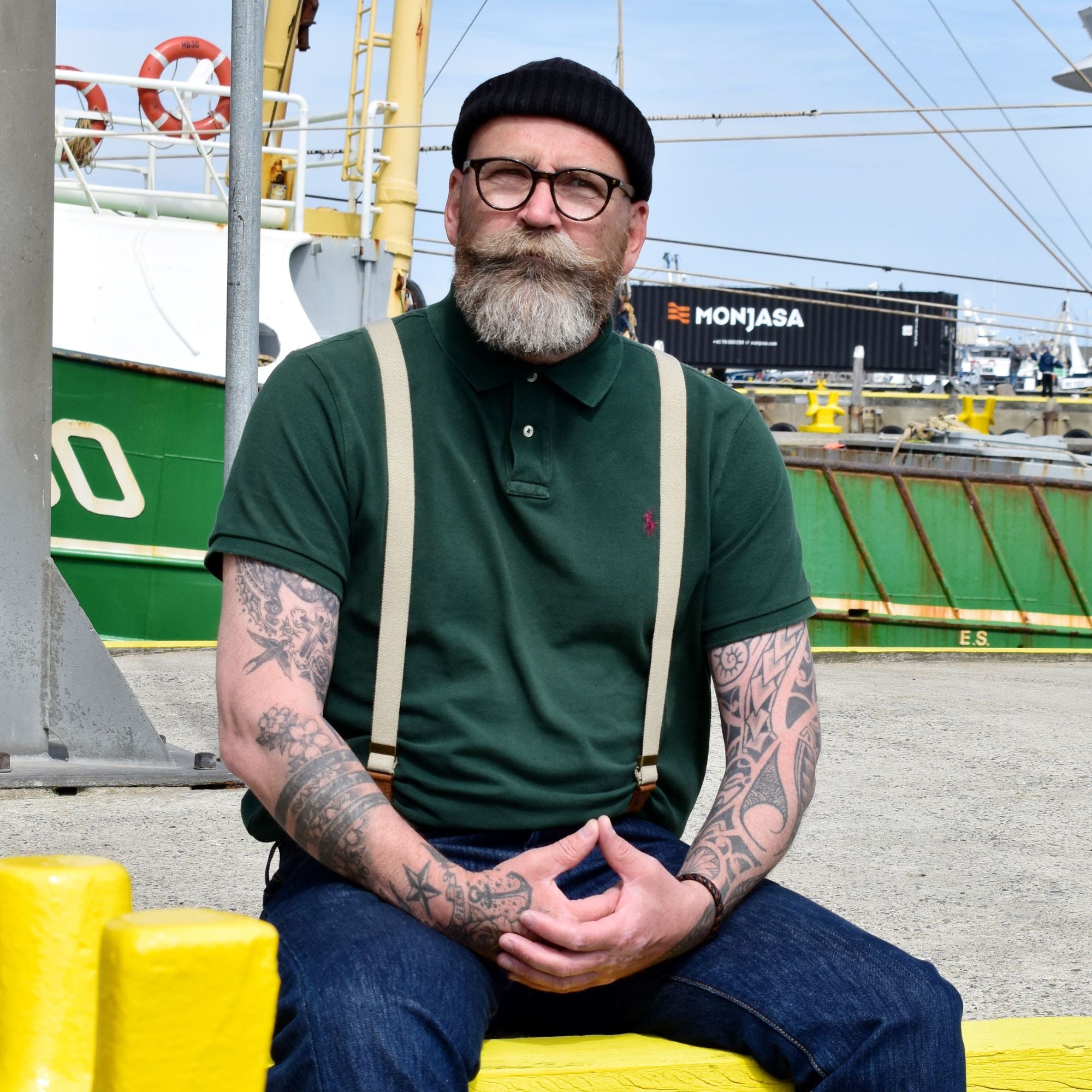 Bearded man is sitting at a dock wearing a beanie, glasses, green shirt and sand colored Wiseguy Original Suspenders