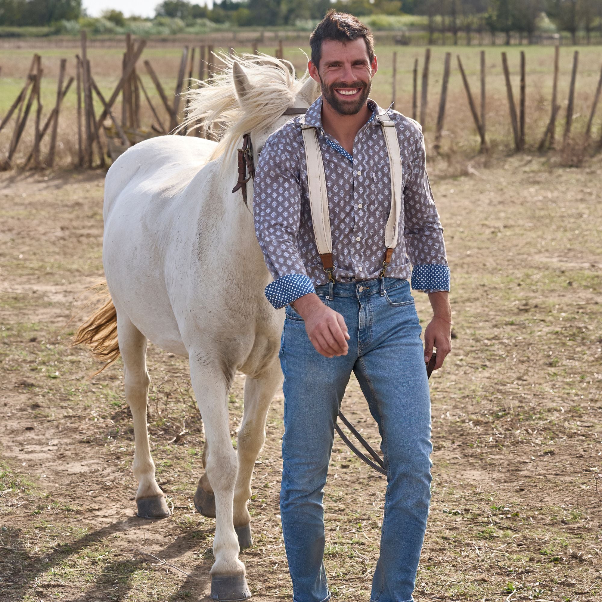 Man wearing white canvas suspenders and a pattered shirt. He is standing in a field with a white horse behing him.