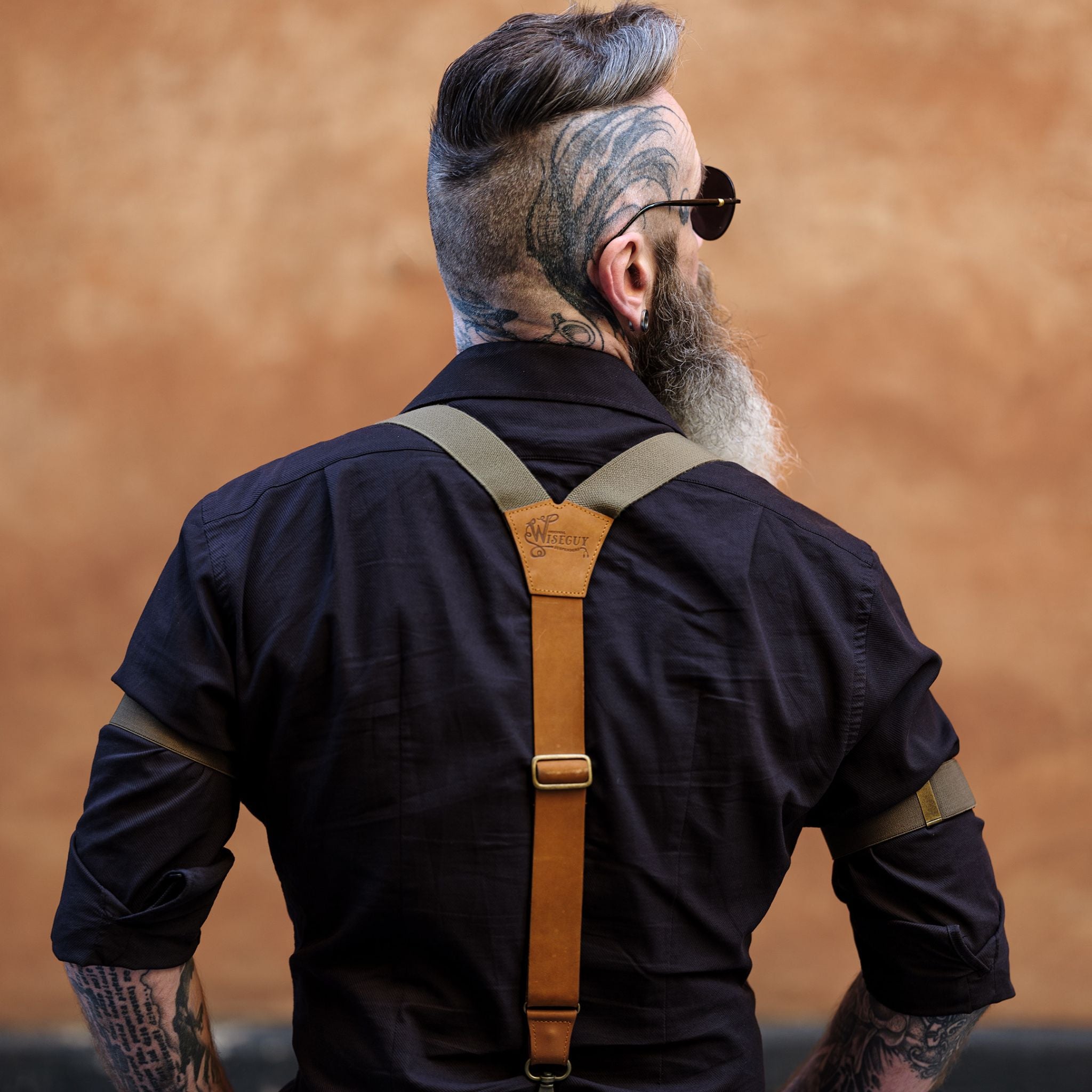 Picture of man from the back wearing a black shirt and camel brown leather suspenders with army green elastic. He is also wearing sleeve garters.