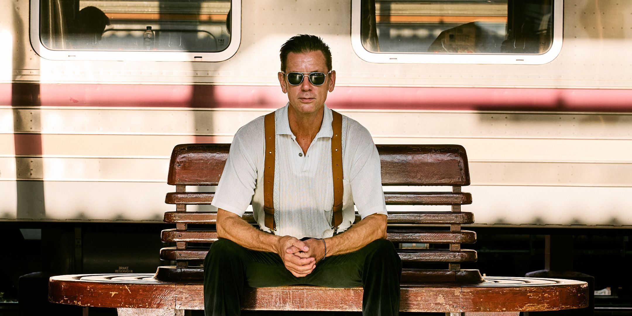 Man Sitting on Train bench wearing sunglasses leather camel colored suspenders and a white tshirt