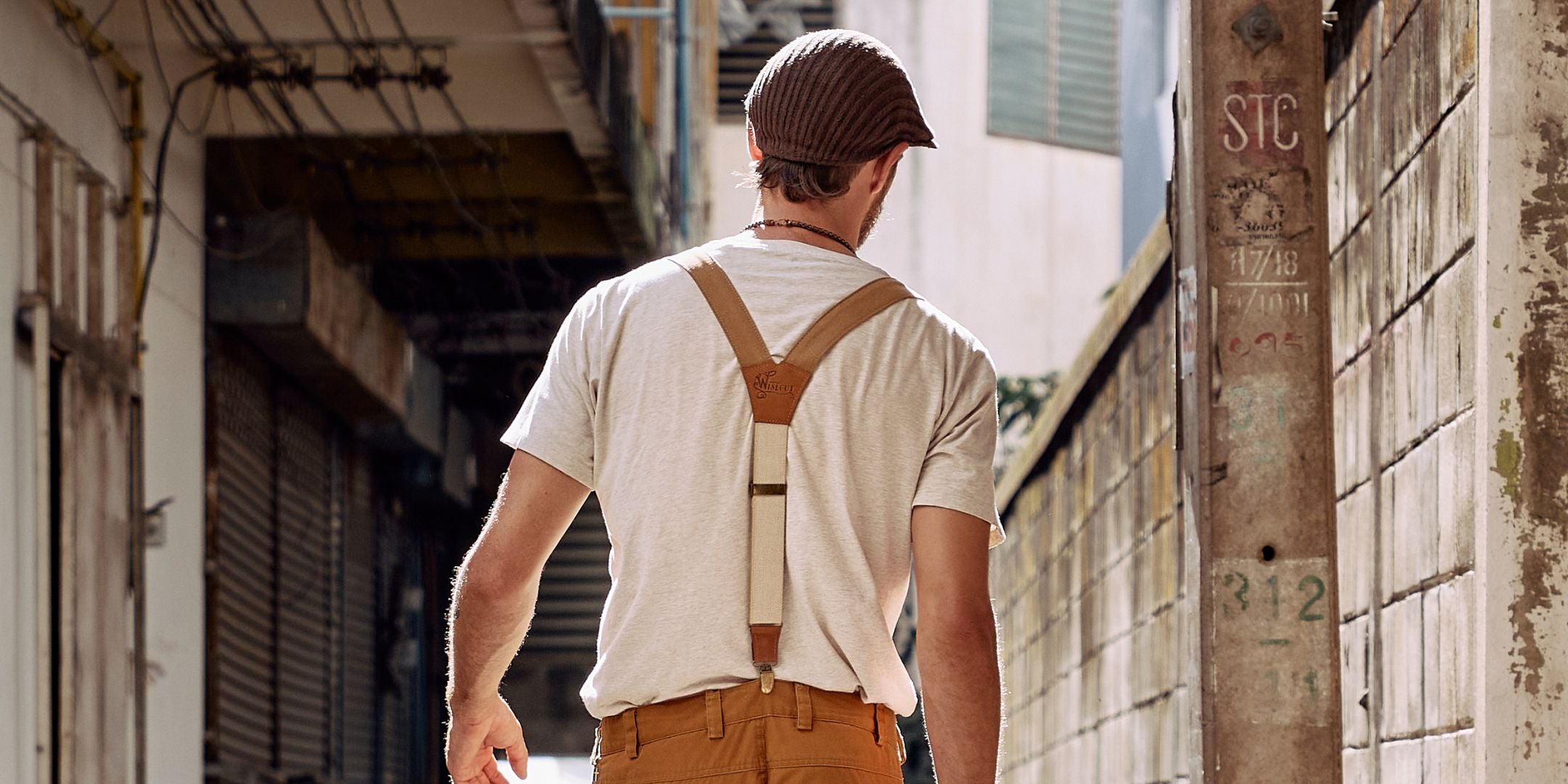 Man walking in street wearing white shirt duo tone colored suspenders and mustard colored pants he is wearing a flat cap